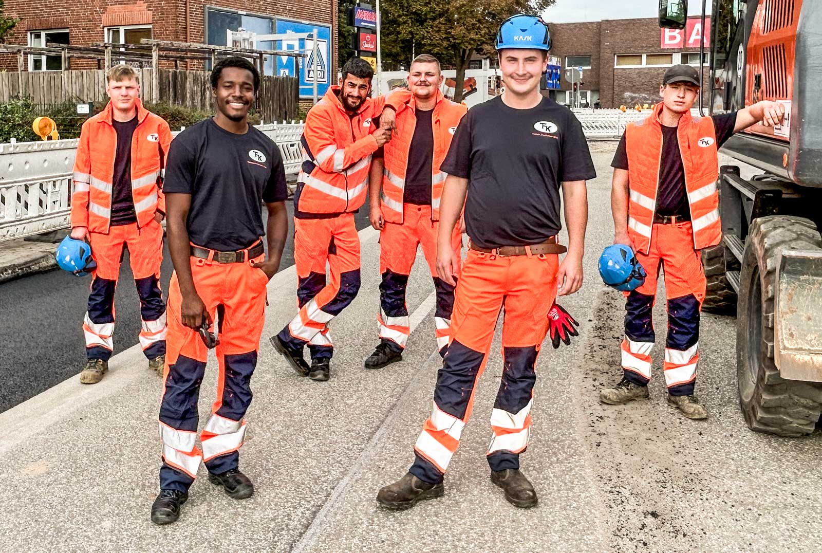 Junges Team in Fasthuber-Arbeitskleidung auf einer Straßenbaustelle neben dem Bagger