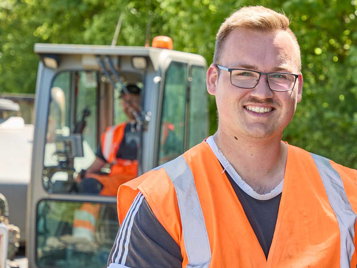 Lächelnde Fachkraft in Warnweste vor einer Baumaschine
