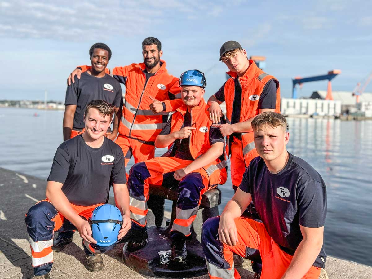 Junges Team in Fasthuber-Arbeitskleidung am Hafen, im Hintergrund Werftkräne