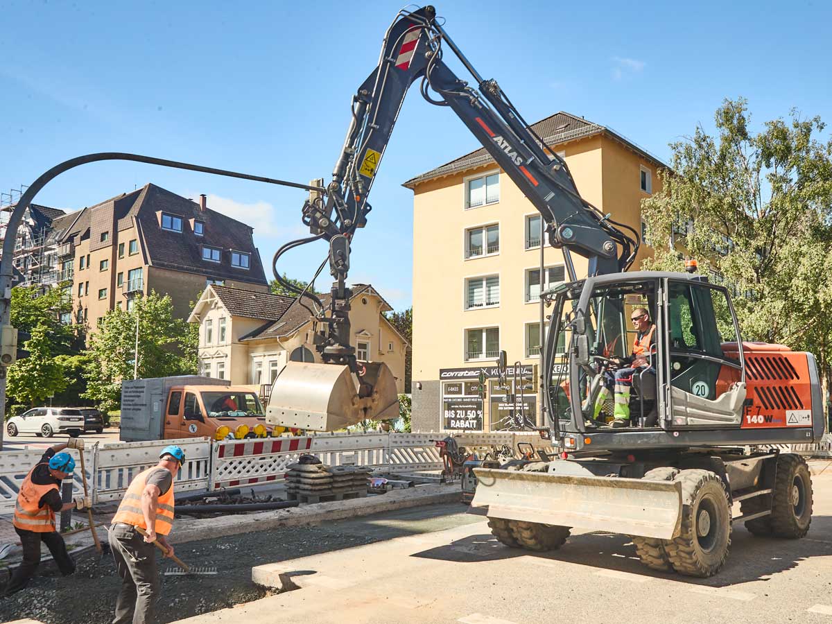 Mobilbagger auf einer Straßenbaustelle, davor zwei Fachkräfte beim Abziehen des Planums